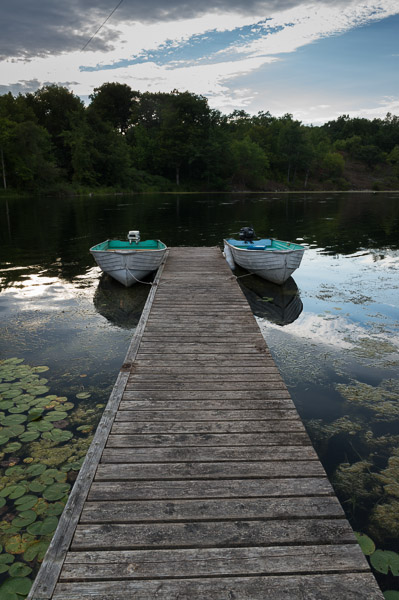 A photo of a lake with two vintage baots, tethered to a long, wooden plank. Behind the boats is lake water, and a bank of the lake with trees.