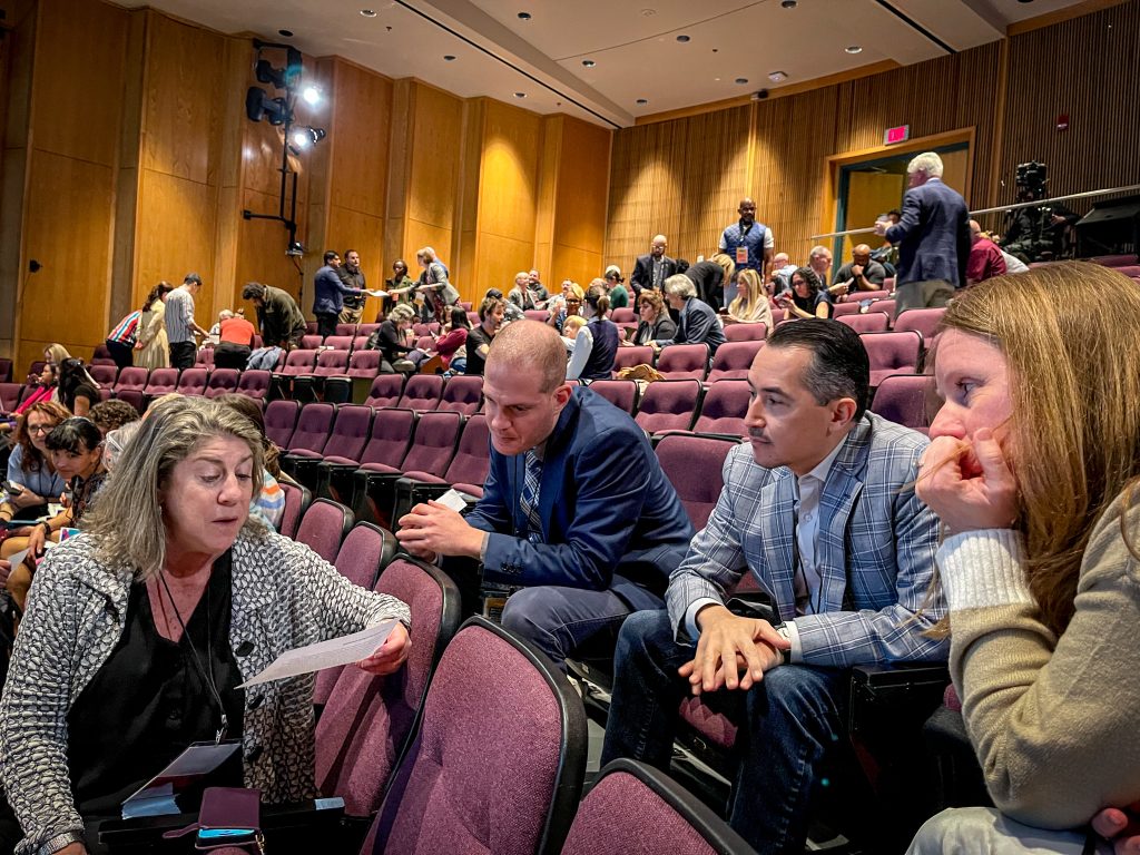 Four individuals discussing in a group,with several additional groups throughout a theater style room.