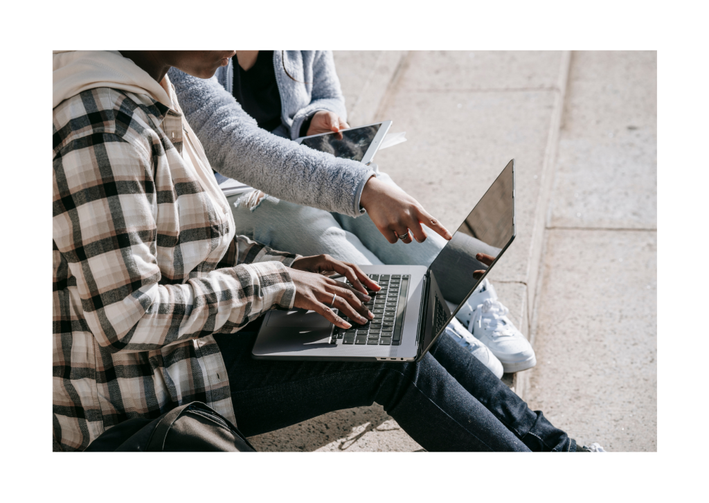 Two students are sitting outside, one is pointing to a laptop screen in a helpful manner.