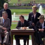 President George Bush sitting at a table on a lawn, surrounded by two individuals in wheelchairs and two others. Bush is signing something on the table.